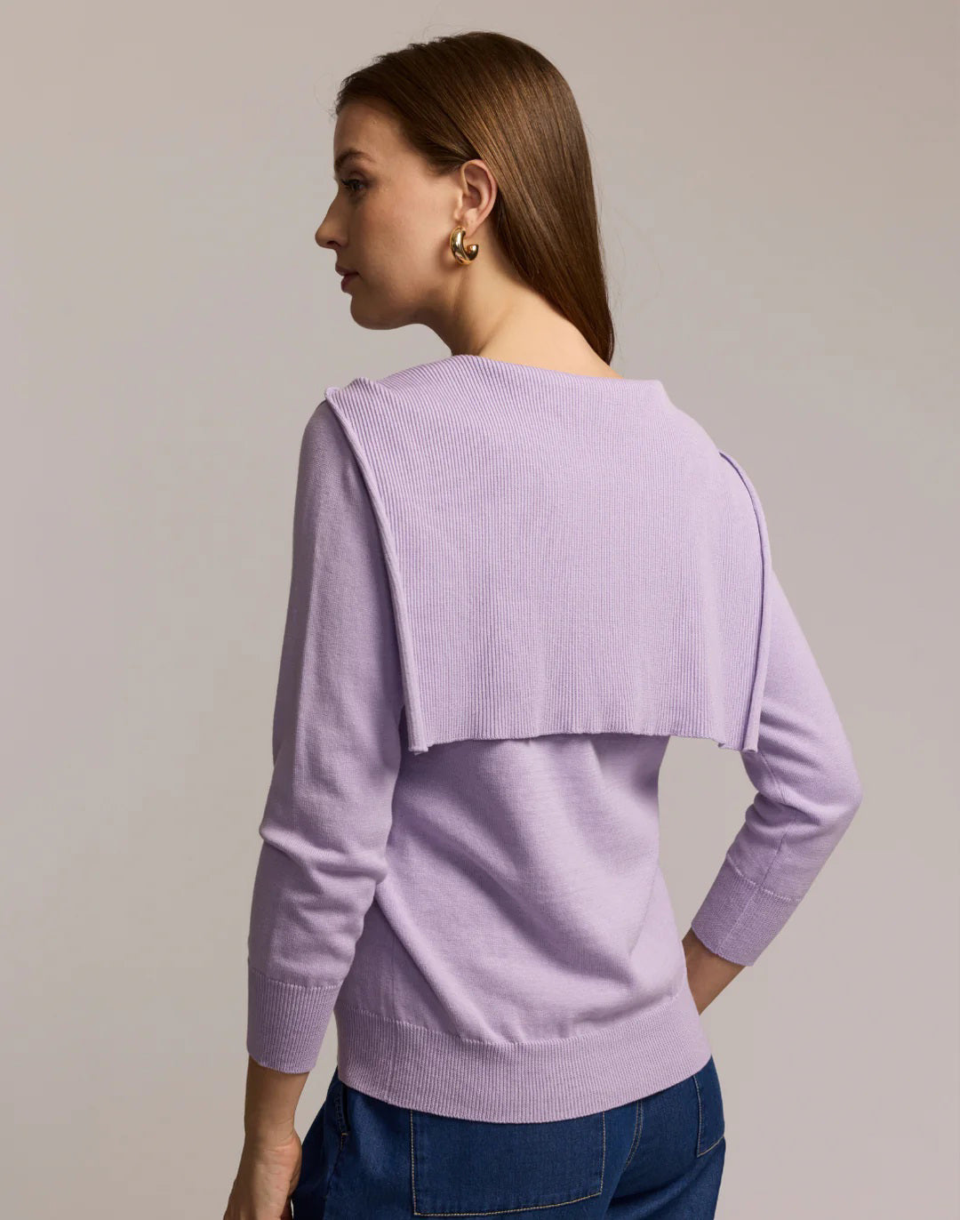 A woman with long brown hair and gold hoop earrings wears blue jeans and a light purple HINSON WU Sloane 3/4 Sleeve Tie Shoulder Cotton Silk Sweater, with a matching sweater draped over her shoulders and tied in front, looking to the side.