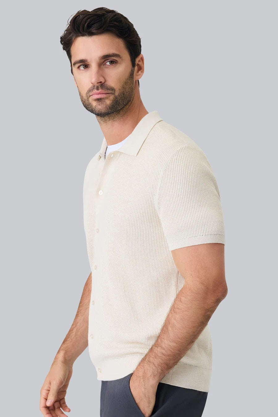 A man with dark hair and a short beard models the MACK WELDON Tech Linen Button Up Sweater Polo in Latte Heather, standing hands-in-pockets against a white background and looking effortlessly stylish and comfortable.