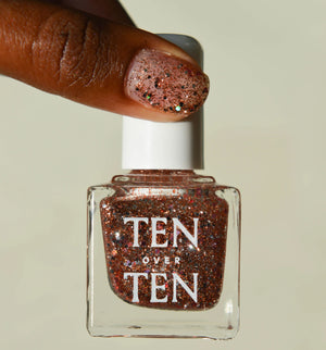A glass bottle of TENOVERTEN - NAIL POLISH from the brand TEN OVER TEN, featuring a white cap and a dusty pink nail color. The front label displays "TEN OVER TEN" in white uppercase letters. This vegan, eight-free formula ensures both beauty and peace of mind. The background is plain and light-colored.