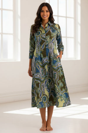A woman stands smiling against a plain beige background, wearing the HINSON WU Martina 3/4 Sleeve Opulent Paisley Print Dress—a long-sleeve, collared, button-down dress featuring a green, blue, and brown paisley pattern and crafted from luxurious Italian polyester fabric for a fluid drape and silky hand feel.