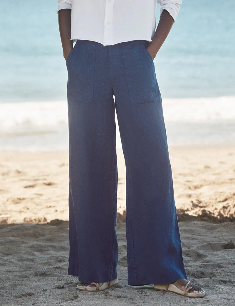 A person stands on a sandy beach wearing a white long-sleeve shirt, FRANK &amp; EILEEN MADELINE Patch Pocket Pull On Wide Leg Linen Pant in Navy, and light-colored sandals. The ocean is visible in the background.