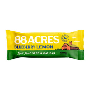 An 88 ACRES - BLUEBERRY LEMON SEED OAT BAR rests in its packaging on a white wooden surface, displaying images of blueberries and lemons. The gluten-free, vegan bar from 88 ACRES is nut-free, nutritious, and delicious.