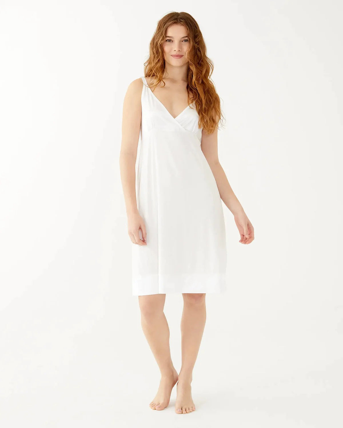 A woman with long wavy hair wears the sleeveless, knee-length MERSEA - MOON DANCER PAJAMA DRESS and smiles gently at the camera, barefoot against a plain white background—a perfect bridal gift.