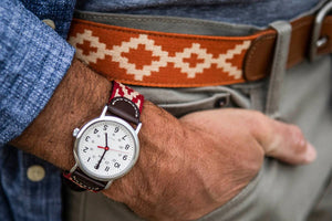A man sporting a checkered shirt and the LA MATERA - TALAMPAYA WOVEN BELT stands by a classic teal car, hand on the door handle, ready to enter. The sunny scene unfolds in a residential area.