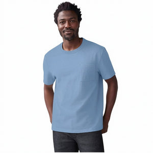 A man with short curly hair wears the MACK WELDON Heavyweight Slub Cotton Pocket T-Shirt in Dean Blue with dark pants, standing against a plain white background.