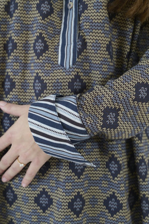 A person with long brown hair, seen from behind, wears the STORIATIPIC MARGOT BLOUSE IN BLUE by STORIATIPIC—a patterned cotton shirt featuring a mix of vertical stripes and diamond shapes in blue and beige tones—paired with blue jeans. The blouse is suitable for a gentle machine wash at 30 degrees.