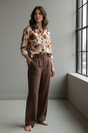 A woman with wavy brown hair stands barefoot indoors, wearing brown wide-leg pants and the HARTFORD HATFORD - CINEMA SHIRT GEOMETRIC PRINT—a loose-fit shirt featuring an abstract orange, beige, and gray pattern—as she gazes out a large window.