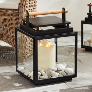 The NAPA HOME AND GARDEN ADWIN OUTDOOR LANTERN features a unique black metal and glass design with a teak handle. Inside, an unlit large white candle enhances its simple and modern appeal.