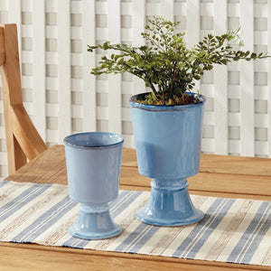 Two NAPA HOME AND GARDEN - BLUE KELCY FOOTED CACHEPOTS, one with a leafy plant and one empty, rest on a wooden table with a blue and white striped placemat, with a white lattice and wooden chair in the background.