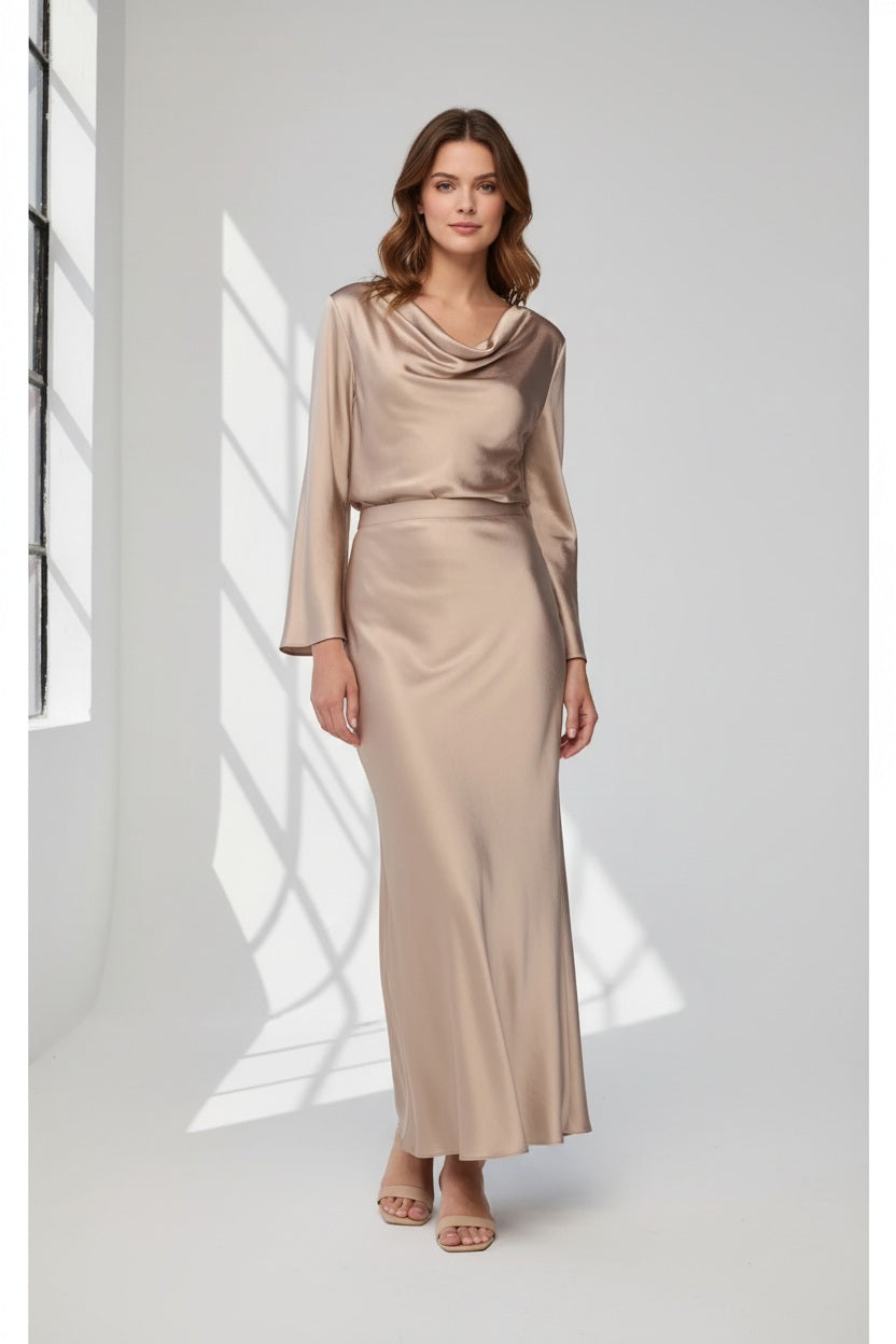 A woman with long blonde hair poses against a plain light gray background, wearing the MILVA SATIN BLOUSE IN SAND by MILVA—a shiny, champagne-beige long-sleeve blouse featuring a chic cowl neckline—paired with a matching skirt and accessorized with bracelets on her left wrist.