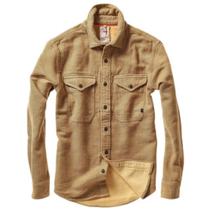 Dressed in the RELWEN Utility Workshirt in cornmeal herringbone layered over a blue and white striped shirt and paired with green pants, the individual stands with relaxed hands by their sides, capturing the effortless sophistication of luxury workwear. The person’s head is partially out of frame.