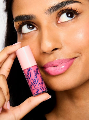 A woman with glossy pink lips holds a tube of LANOLIPS Hyaluronic Lip Oil in Raspberry near her face, looking upward with a slight smile, showcasing the non-sticky lip oil’s effortless lip hydration.