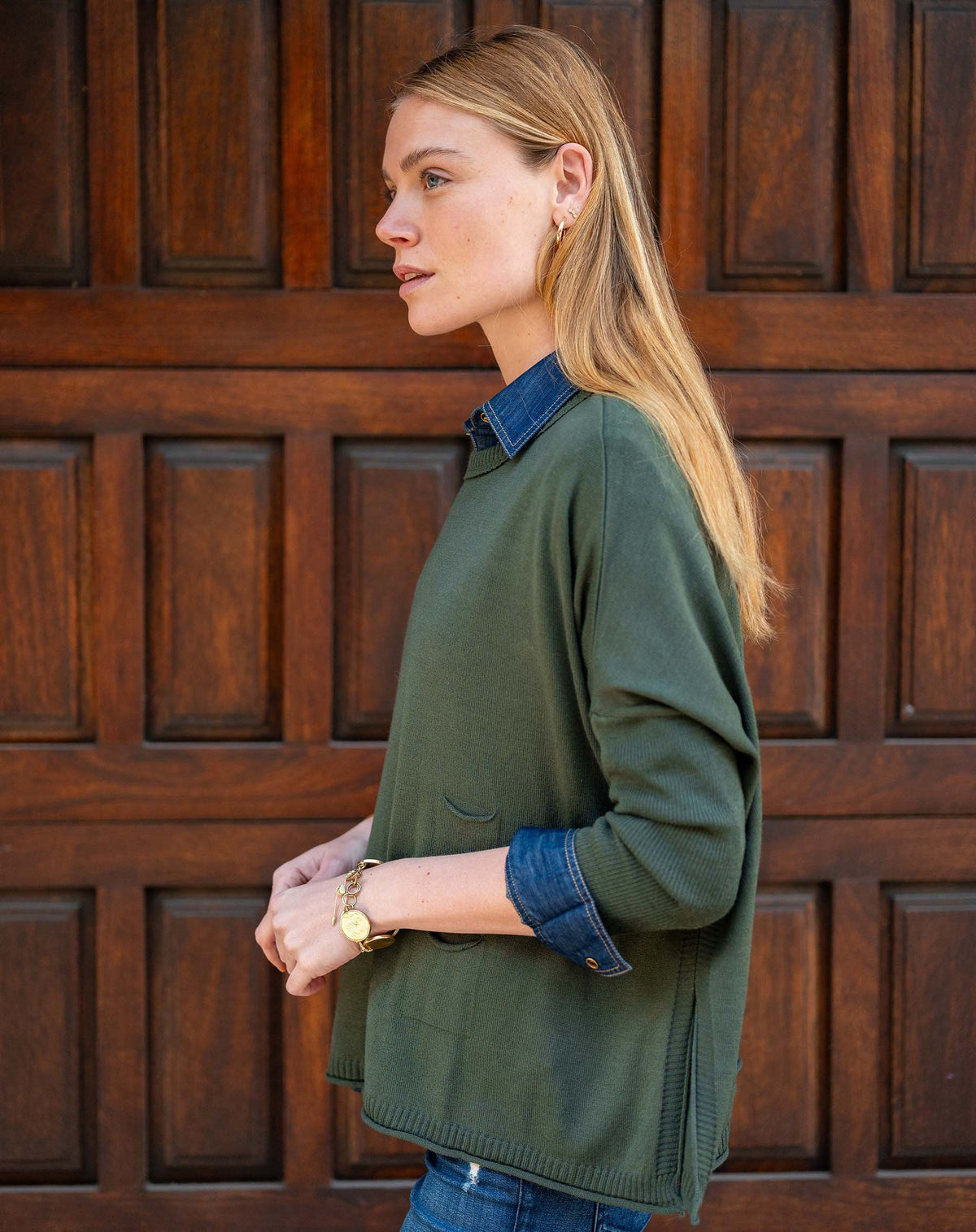 A woman with light brown hair stands before a wooden door, wearing the MERSEA - CATALINA CREWNECK SWEATER IN DEEP EMERALD over a denim shirt and blue jeans, gazing to the side with hands in her pockets and a relaxed expression.