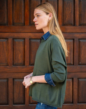 A woman with light brown hair stands before a wooden door, wearing the MERSEA - CATALINA CREWNECK SWEATER IN DEEP EMERALD over a denim shirt and blue jeans, gazing to the side with hands in her pockets and a relaxed expression.
