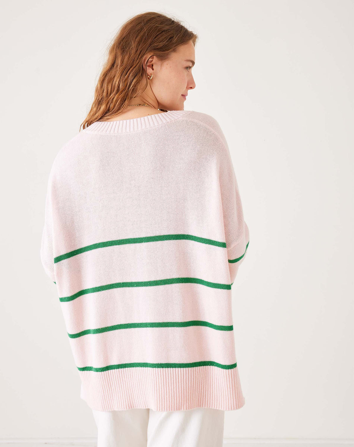A woman wears the MERSEA - MONTAUK V-NECK SWEATER by MERSEA, a white oversized soft knit with blue and green striped collar, cuffs, and hem, ribbed v-neckline, styled over denim shorts and a light pink shirt.