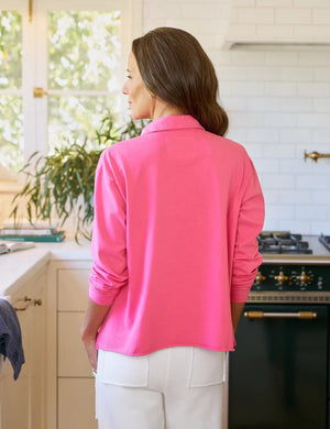A woman with long brown hair stands in a sunlit kitchen, wearing the FRANK & EILEEN Patrick Popover Henley in Lotus and white pants. Light pours through a window adorned with plants, capturing California Heritage Collection vibes.