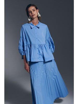 A woman with straight, light brown hair wears the MILVA MI - COTTON BLOUSE IN AZZURO BLUE by MILVA, featuring voluminous sleeves and a peplum hem, paired with bold white earrings against a plain gray background.