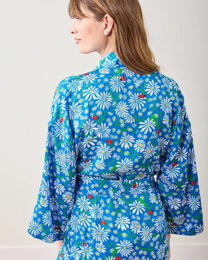 A woman stands barefoot on a wooden floor wearing the PRINTFRESH Satin Mini Robe Lucky Daisy. The blue floral pattern drapes elegantly with long sleeves and a waist tie as she gazes sideways against a light wall, showcasing relaxed elegance and sustainable style.