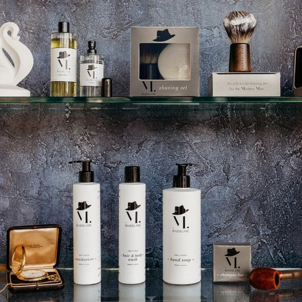 The BAUDELAIRE - M.B. SHAVING SET by BAUDELAIRE includes a luxurious shaving brush and premium shaving soap, presented in an elegant box featuring a delightful hat illustration and “shaving set” printed on the front. Displayed outdoors atop lush moss, this refined ensemble adds classic sophistication to your grooming routine.