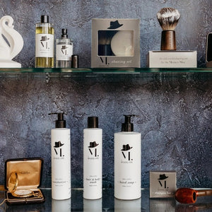 The BAUDELAIRE - M.B. SHAVING SET by BAUDELAIRE includes a luxurious shaving brush and premium shaving soap, presented in an elegant box featuring a delightful hat illustration and “shaving set” printed on the front. Displayed outdoors atop lush moss, this refined ensemble adds classic sophistication to your grooming routine.