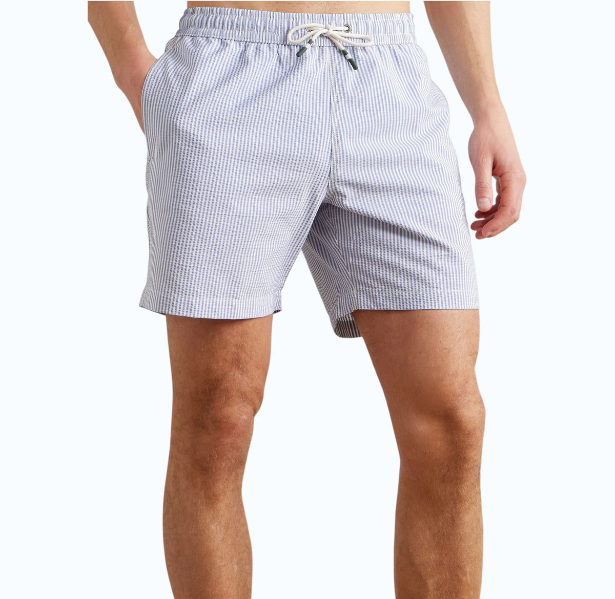 A person wears HARTFORD - SEERSUCKER SWIM TRUNKS IN INDIGO with a drawstring waist, side pockets, and a quick-drying seersucker fabric. The mid-thigh trunks are shown against a plain background, displaying only the torso and legs.