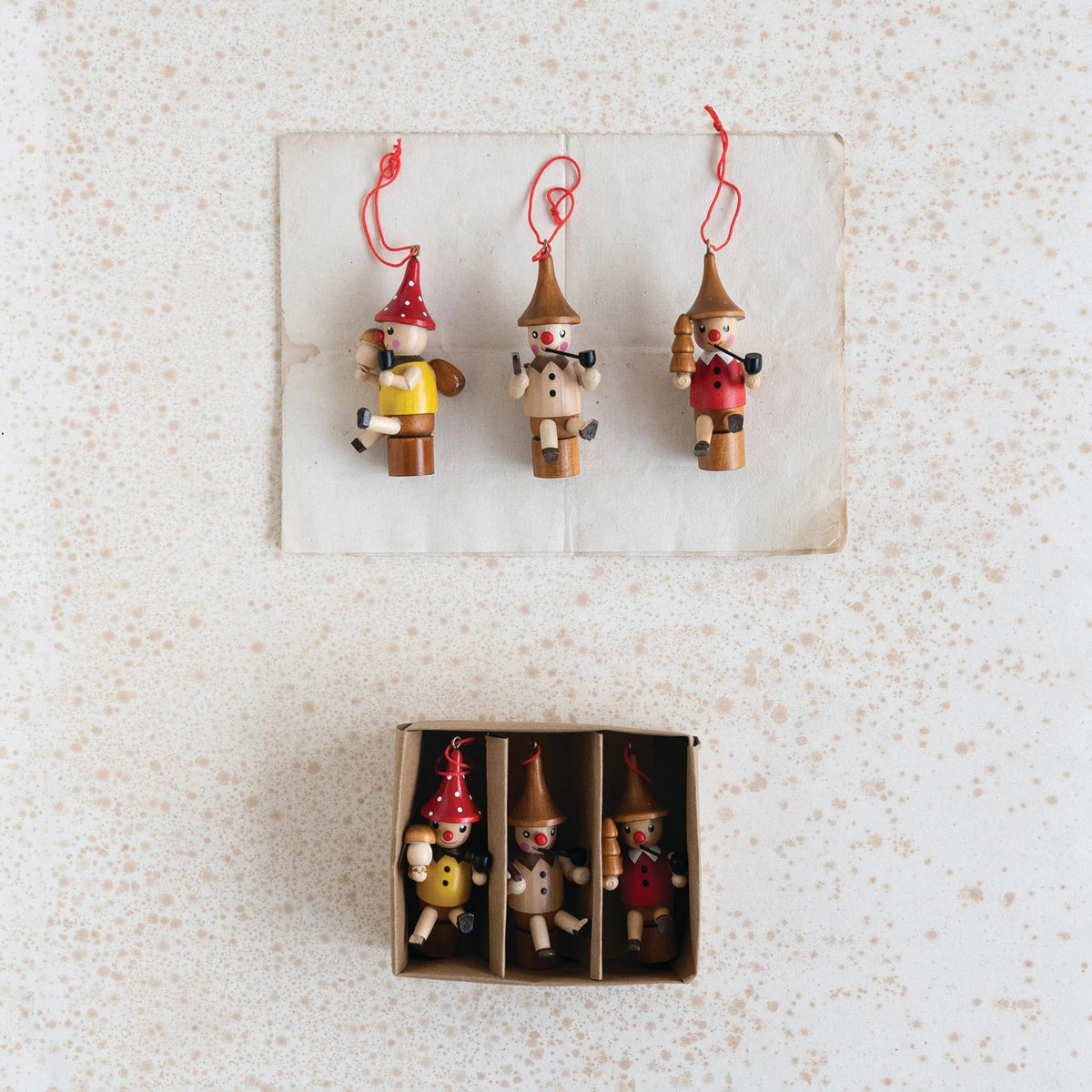 Three CREATIVE COOP WOOD WOODSMEN ORNAMENTS with red hats hang above a boxed set of four similar multi-colored figures, each in its own compartment, all displayed on a speckled white surface.