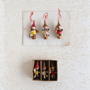 Three CREATIVE COOP WOOD WOODSMEN ORNAMENTS with red hats hang above a boxed set of four similar multi-colored figures, each in its own compartment, all displayed on a speckled white surface.