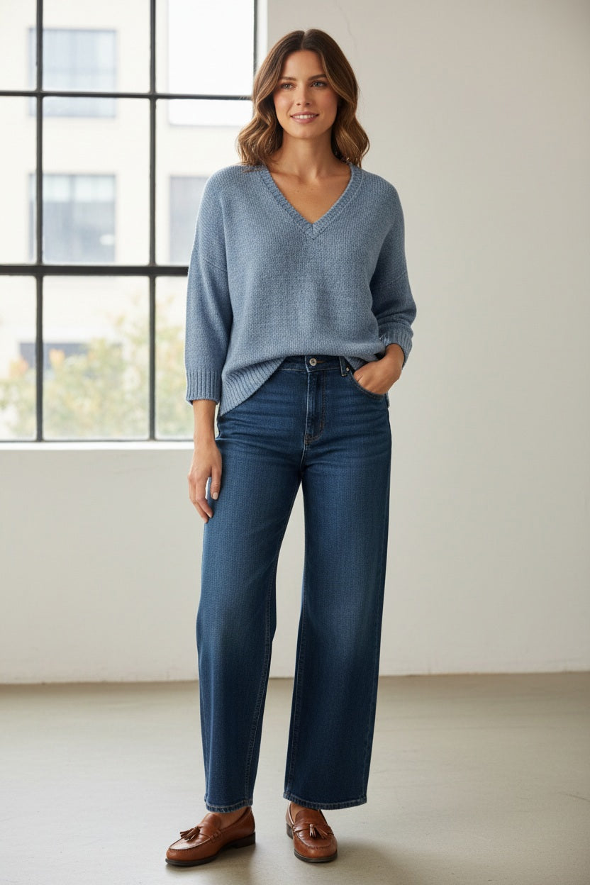 A smiling woman with long blonde hair wears the BASE MILANO - V NECK COTTON SWEATER, a light blue, short-sleeved, relaxed fit knit by BASE MILANO, paired with blue jeans. She stands against a plain background, hand on her chin.