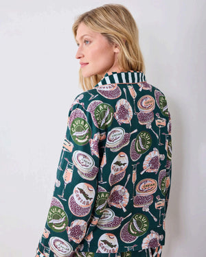 A woman stands barefoot on a wooden floor against a white wall, wearing the PRINTFRESH - Champagne & Caviar Eco Satin Soho Set in Peacock Feather by PRINTFRESH, a matching dark loungewear set adorned with colorful floral and fruit motifs and finished with checkered cuffs.