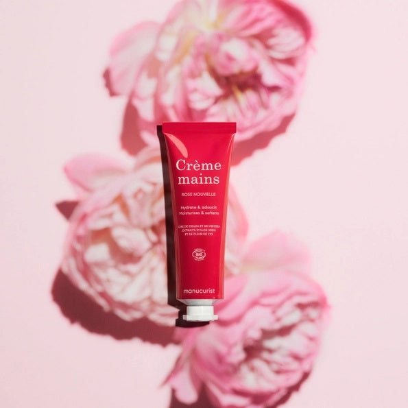 With perfectly manicured nails, a person applies MANUCURIST - COSMOS ORGANIC HAND CREAM, ROSE NOUVELLE from MANUCURIST INC to the back of their hand against a soft pink background, emphasizing the importance of caring for hands and nails.