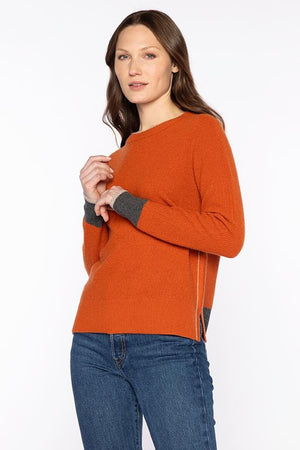 A woman with long brown hair models the KINROSS Tipped Thermal Cashmere Crewneck Sweater in burnt orange with dark gray cuffs, paired with blue jeans, standing against a plain white background.