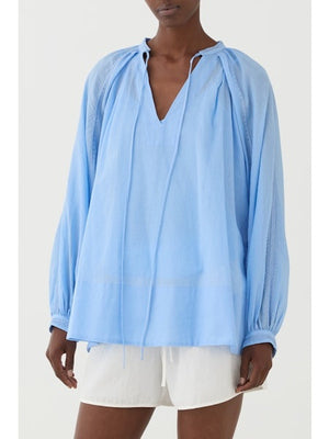 A person wearing the ELEH - BLOUSE WITH EMBRIODERY by ELEH, a loose, light blue peasant-style blouse with long sleeves and tie details, paired with white shorts, stands against a plain light background.
