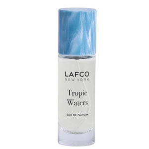 A LAFCO FINE FRAGRANCE, TROPIC WATERS 10ML box stands upright in a halved coconut, water splashing around it against a neutral background—an evocative display for this long-lasting scent inspired by the essence of coconut water.
