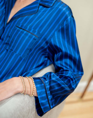 The MERSEA - Carousel Stripe Satin Sailor Pajama Set in Blue by MERSEA, featuring elegant blue and black stripes with a button-up top and matching pants, embodies luxury sleepwear and is neatly folded on a light blue background.