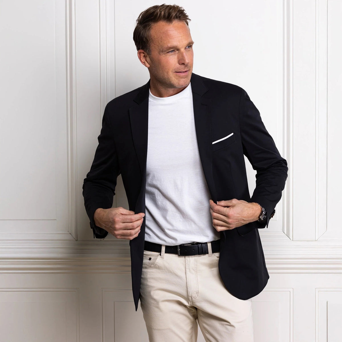A man in the COLLARS AND CO Maverick Performance Blazer in Stealth Black, layered over a white t-shirt and beige pants, stands by a white paneled wall, looking to the side as he adjusts his blazer.