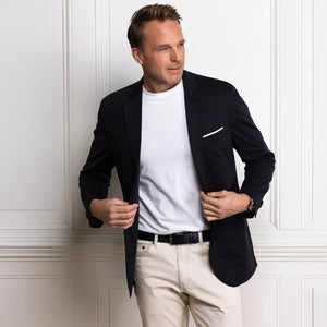 A man in the COLLARS AND CO Maverick Performance Blazer in Stealth Black, layered over a white t-shirt and beige pants, stands by a white paneled wall, looking to the side as he adjusts his blazer.