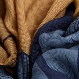 A woman stands against a white background, wearing a cream jacket, beige pants, and INOUI EDITIONS’ Cavale Horse Wool Scarf in Navy—hand-printed in India with a vibrant unicorn design—draped over her neck and shoulder.