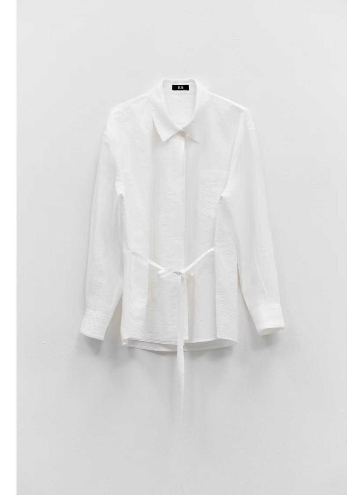 The ELEH LONG SLEEVE SHIRT WITH TIES by ELEH, crafted from a white Italian cotton blend, hangs neatly against a plain white wall. Featuring a collar and waist tie detail, this modern button-down silhouette is perfect for any occasion.