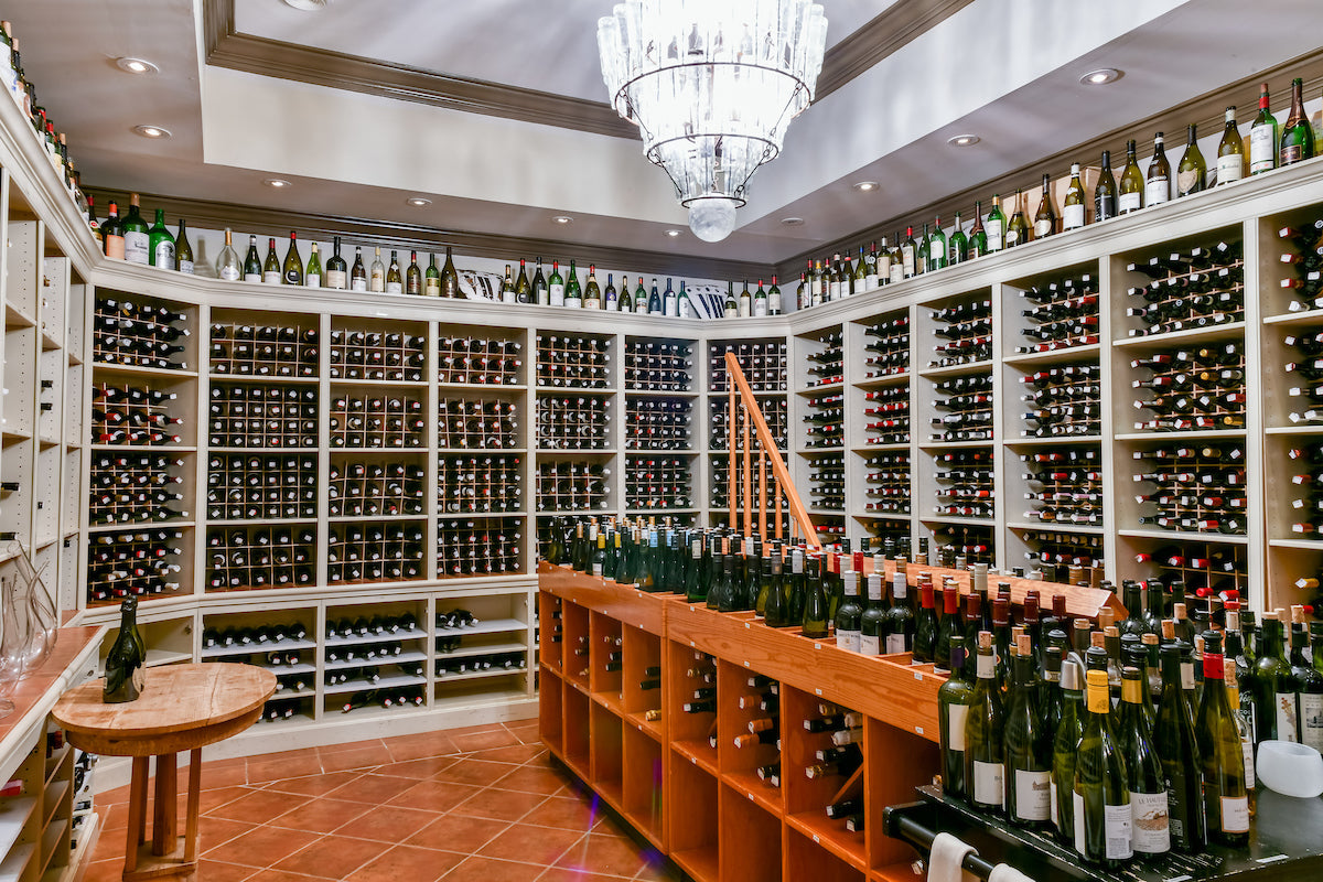 A well-lit wine cellar with white shelves filled with hundreds of wine bottles, a wooden table, a chandelier, and a ladder for reaching high shelves. More bottles are displayed on the countertop and along the top shelves.