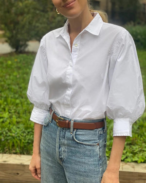 A pink button-front shirt, THE LITTLE PROJECT's ESME SHIRT IN STRAWBERRY, is displayed flat on a white surface. It has a straight cut with one visible button near the collar and a "100% Linen" tag featuring the brand logo and size label.