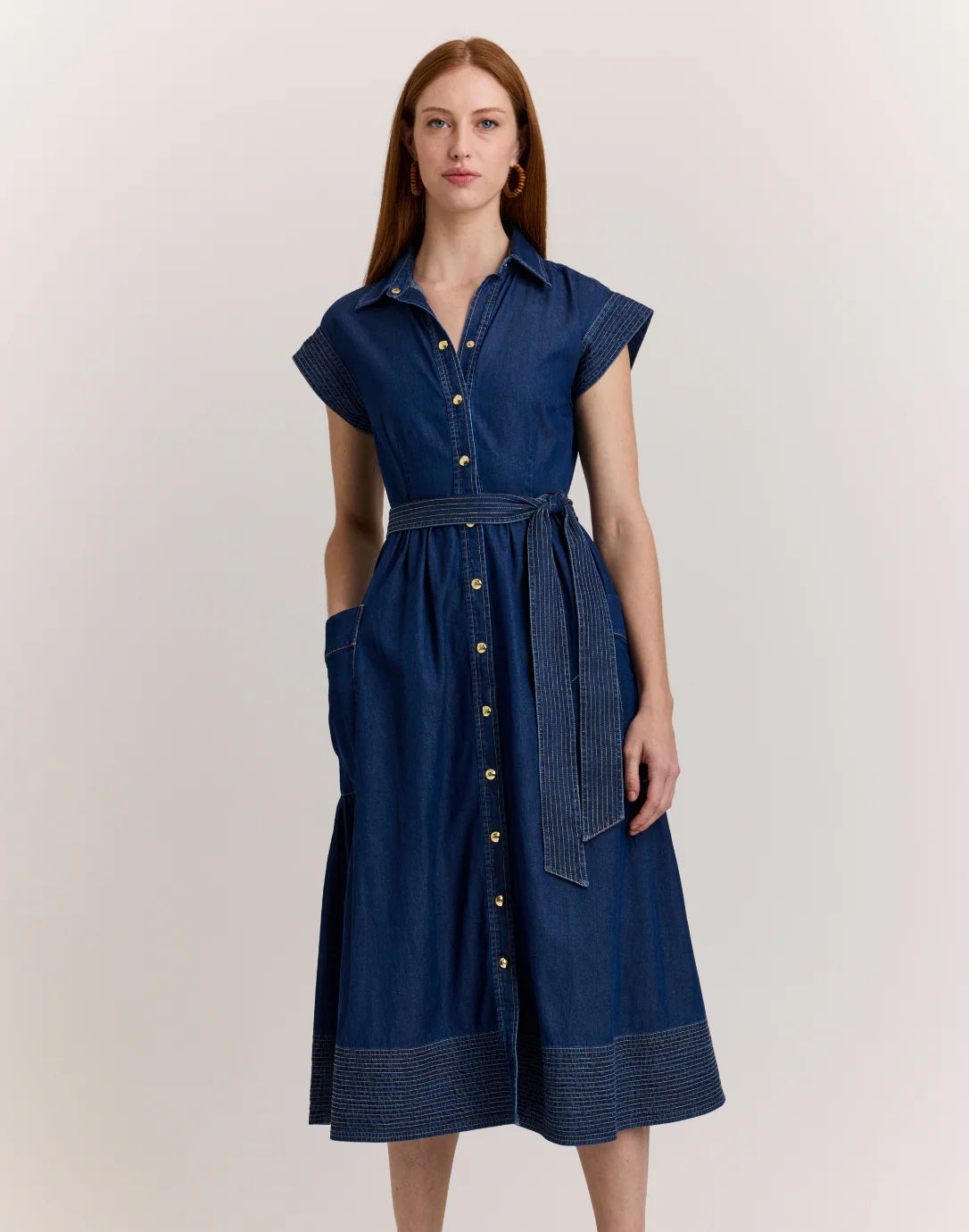 A woman with long auburn hair wears the HINSON WU ANNABELLE Short Sleeve Dark Wash Denim Dress, featuring a belted waist, as she stands facing away from the camera against a light background.