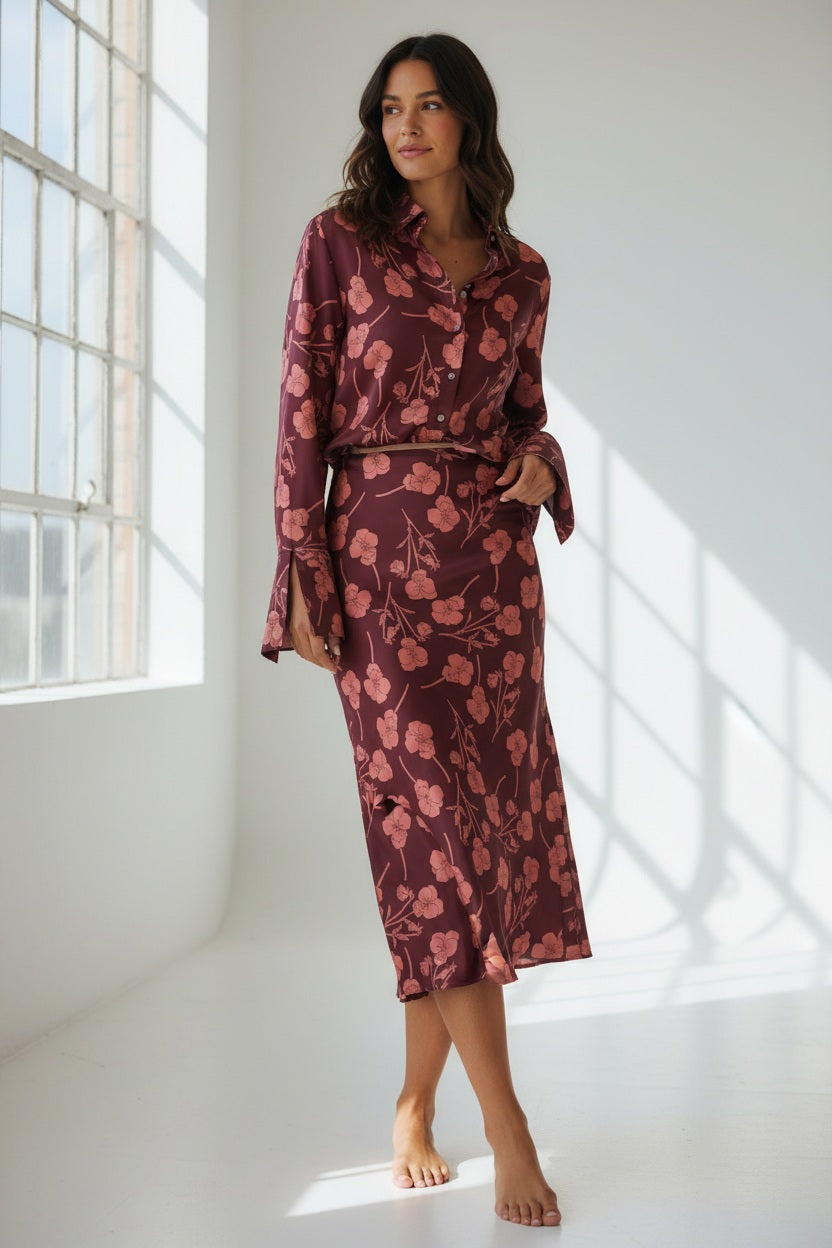 A woman stands barefoot in a sunlit room, wearing the MIRTO 1956 - FLORAL SILKY MIDI SKIRT paired with a long-sleeve, burgundy floral blouse. She gazes to the side with a relaxed expression, her hands tucked into her skirt pockets.