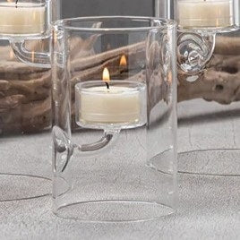 ZODAX SUSPENDED GLASS TEALIGHT HOLDER - EXTRA SMALL displays red tealights burning in clear glass holders of varying heights, set on a white surface with pine branches, berries, and a pinecone against a deep red background.