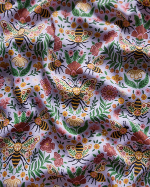 A person wearing the PRINTFRESH - BEE TAPESTRY ECO SATIN SOHO SET IN LAVENDER, featuring vintage-inspired bee and floral vine prints, holding a flower behind their back in a garden.