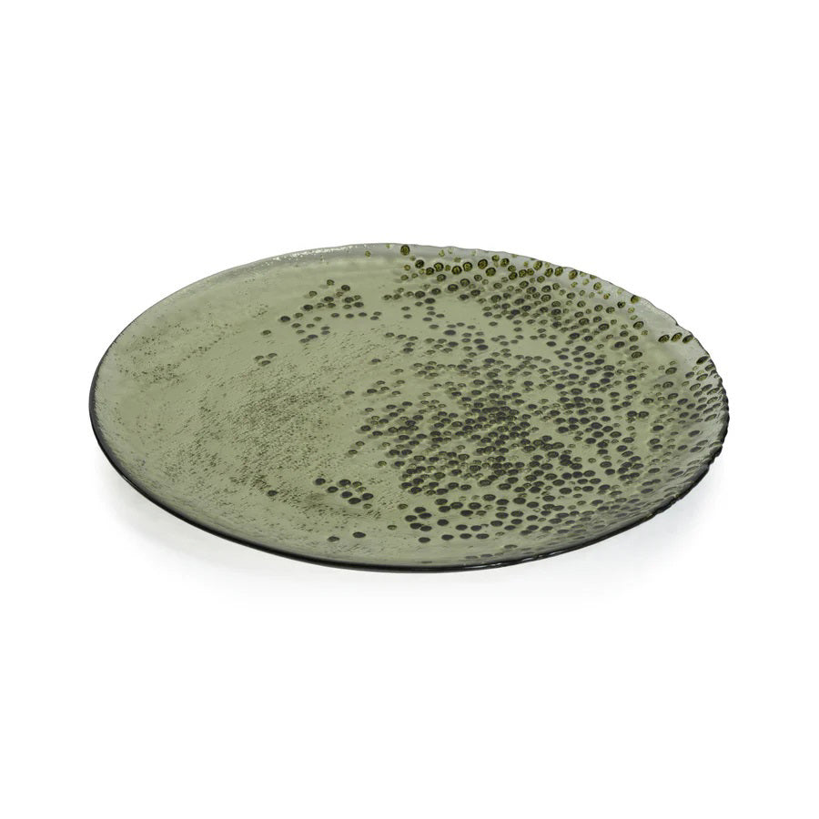 The DOTTED GLASS PLATE 11" by ZODAX is a round ceramic plate with an 11-inch diameter and a green glazed finish. Its surface displays a pattern of small, raised dots that are concentrated on the right side and gradually spread towards the center. The edges of this meticulously crafted plate are slightly uneven.