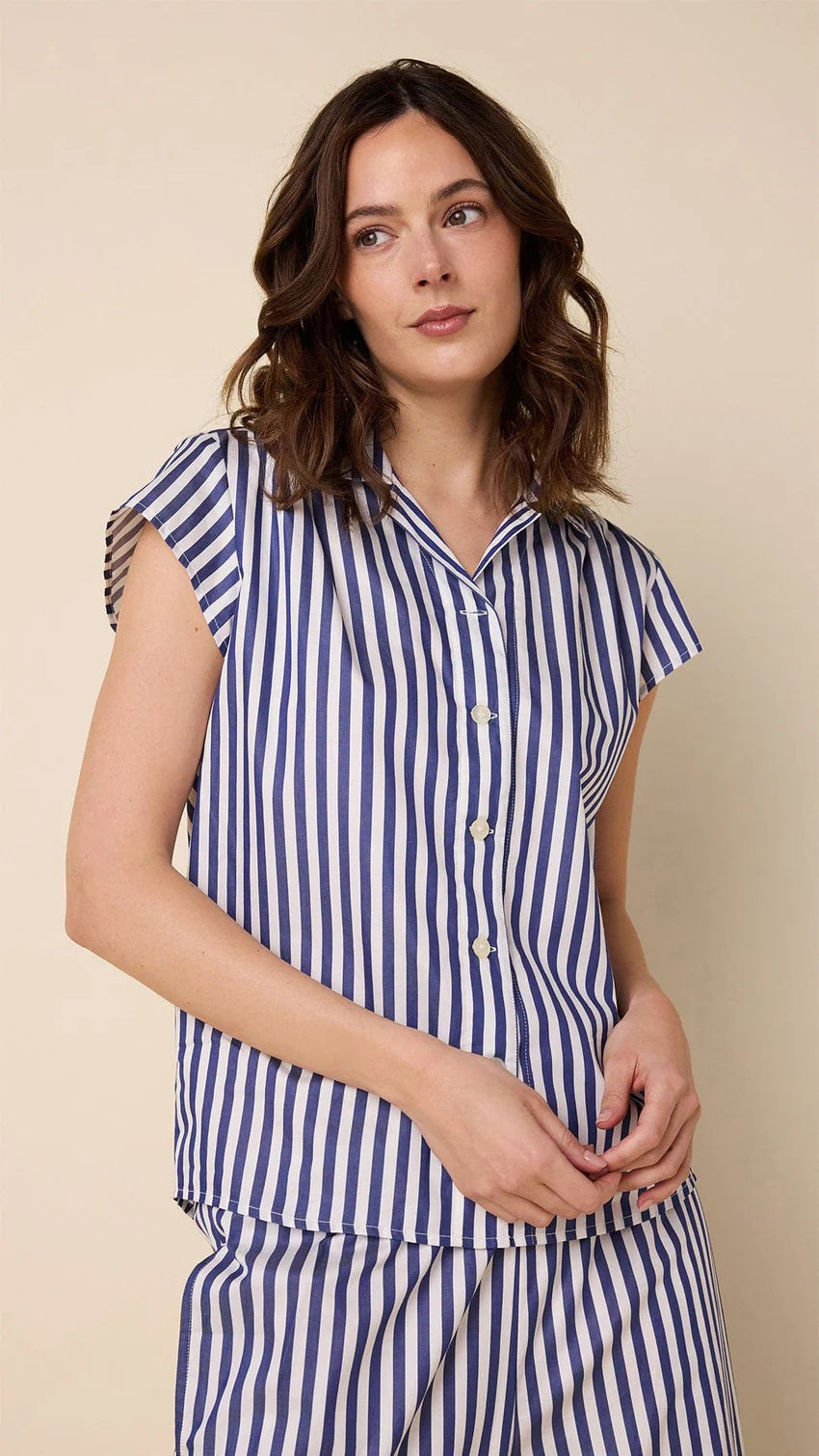 A woman wears CAT&#39;S PAJAMAS - CLASSIC STRIPE LUXE PIMA CAPRI PAJAMAS with blue and white vertical stripes, short sleeves, and cropped pants, standing barefoot against a plain beige background.