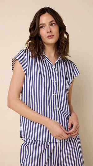 A woman wears CAT'S PAJAMAS - CLASSIC STRIPE LUXE PIMA CAPRI PAJAMAS with blue and white vertical stripes, short sleeves, and cropped pants, standing barefoot against a plain beige background.