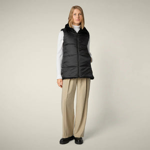 A woman with straight blonde hair wears the SAVE THE DUCK NIVES REVERSIBLE VEST over a white long-sleeve top, beige wide-leg pants, and black shoes, standing against a plain background and highlighting the vest's chic style.