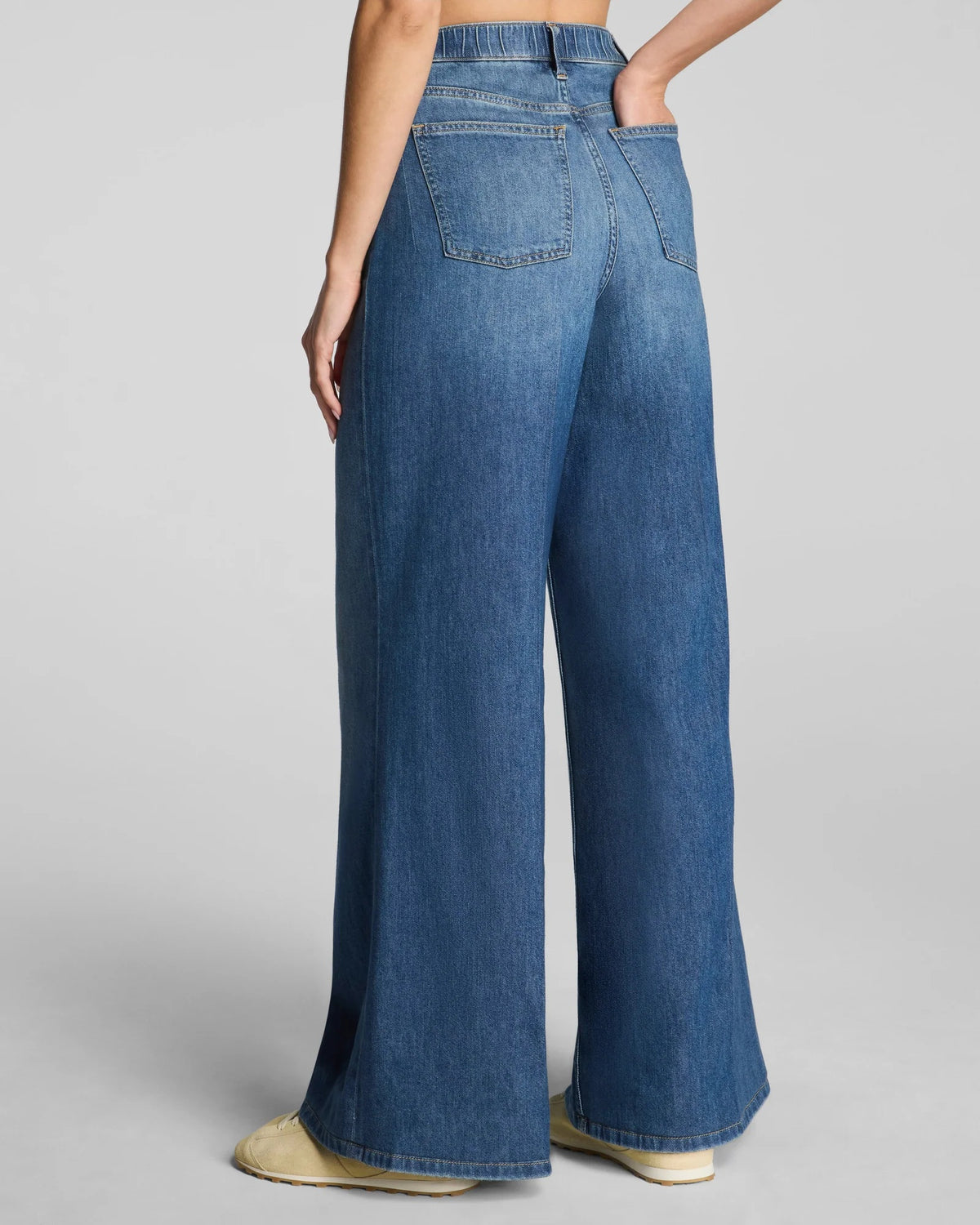 A person shown from mid-torso down against a plain gray background wears the SPANX SPANXSUPERSMOOTH Authentic Lightweight Super Wide Leg Jean in Oceanic paired with beige shoes.