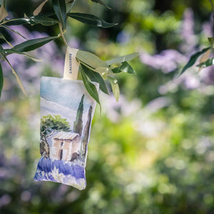 The L'AUGUSTE PROVENCE - FRENCH LAVENDER SACHET FROM PROVENCE is a small handcrafted pouch with a countryside watercolor design, upright shape, "L'AUGUSTE PROVENCE" tag, and dried lavender pieces at its base.